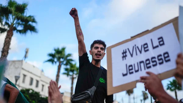 Un jeune homme le poing levé au milieu d'une foule lors d'une manifestation du mouvement GenZ 212 au Maroc, derrière une banderole portant l'inscription « #Vive La Jeunesse ».