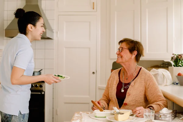 Une auxiliaire de vie en chemise bleu clair, souriante et tenant une assiette dans une cuisine, face à une femme âgée aux cheveux courts et lunettes assise à table qui lui sourit.