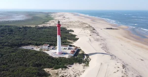 Vue aérienne du phare de La Coubre à La Tremblade, haut phare rayé rouge et blanc dominant la côte sablonneuse, entouré de végétation verte et d'une longue plage, face à l'océan, menacé par l'érosion.