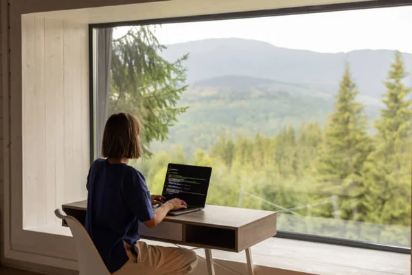 Travailleur en télétravail assis à un bureau en bois devant une grande fenêtre donnant sur un paysage montagneux boisé, écran d'ordinateur portable affichant du code informatique, vêtu d'un t-shirt bleu et de pantalon beige, dans un cadre de campagne.