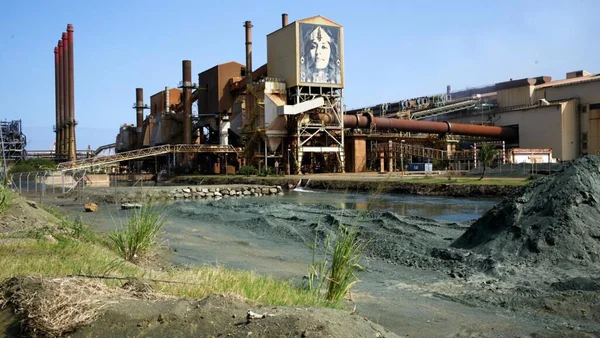 Usine SLN à Nouméa après les émeutes de juin 2024, avec des cheminées fumantes, un grand mur d'une face humaine et des tas de matière sombre derrière l'eau.