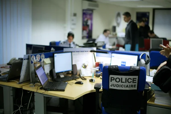 Photographie de l'intérieur d'un bureau de police judiciaire avec une chaise bleue portant le logo 'POLICE JUDICIAIRE' et des cyber-enquêteurs travaillant à des ordinateurs.