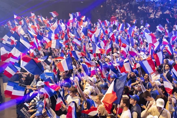 Foule nombreuse dans une salle couverte agitant de nombreux drapeaux français, créant une mer de bleu, blanc et rouge.