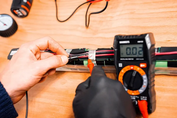 Mains utilisaient un multimètre numérique pour tester la tension d'une batterie de vélo électrique posée sur une étagère en bois, environnement de atelier.