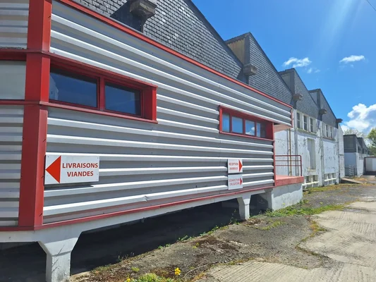 Extérieur de l'abattoir de Vannes, bâtiment à bardage métallique ondulé et liseré rouge, avec panneaux en français indiquant les livraisons de viande et la réception des animaux.