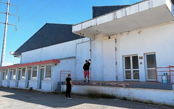 Deux hommes devant l'abattoir du Prat à Vannes : l'un sur le quai de chargement en short rouge et sweat noir, l'autre en tenue noire sur le gravier, devant un bâtiment blanc.