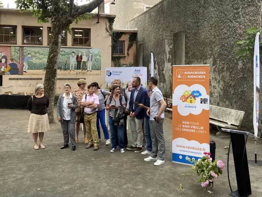 Groupe de personnes debout dans une cour extérieure, à côté d'une banderole d'une association de soutien aux seniors LGBT+, à la Croix-Rousse.