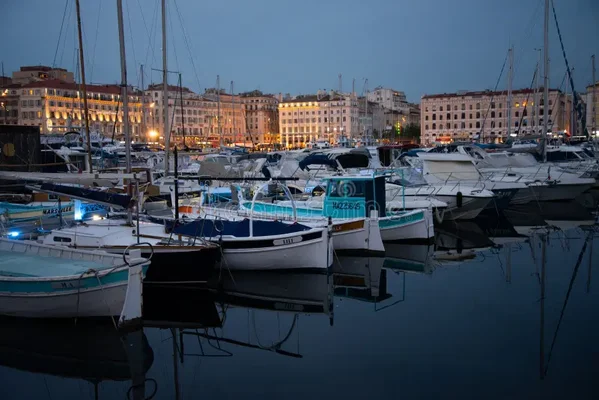 Le Vieux-Port de Marseille au crépuscule : nombreux bateaux amarrés dans une eau calme, reflets des lumières, immeubles du centre historique éclairés en arrière-plan