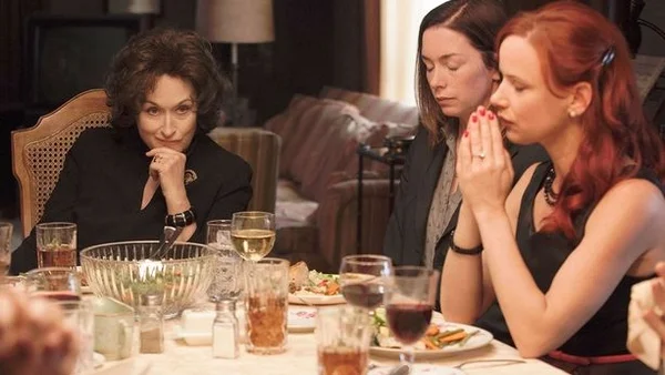 Scène de film : trois femmes attablées lors d'un repas de famille ; l'une, cheveux courts foncés, le menton posé sur la main, face à deux jeunes femmes les mains jointes.