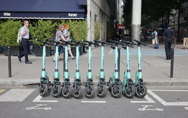 Rangée de trottinettes électriques turquoise stationnées dans une voie réservée à Paris, avec des piétons passant devant un bistrot en arrière-plan.
