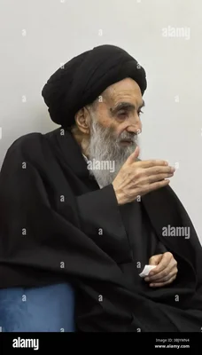 Le grand ayatollah Ali al-Sistani, âgé, barbe blanche, turban noir et robes noires, assis, tenant un papier et faisant un geste de la main.