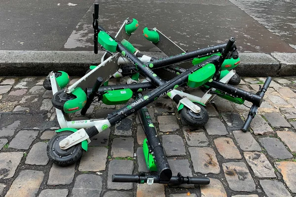 Trottinettes électriques Lime vertes et noires renversées en tas sur une rue pavée mouillée de Paris, au bord d'un trottoir.