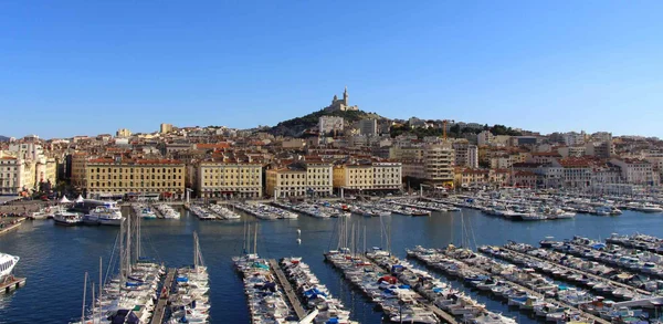 Vue panoramique du Vieux-Port de Marseille sous un ciel bleu dégagé, avec les bateaux amarrés dans le bassin, les immeubles de la ville et la basilique Notre-Dame de la Garde dominant la colline en arrière-plan