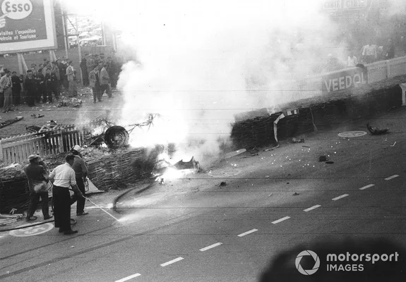 Photographie en noir et blanc de l'épave en feu de la Mercedes 300 SLR de Pierre Levegh lors de l'accident des 24 Heures du Mans, avec des pompiers tentant d'éteindre les flammes et des spectateurs derrière une barrière.