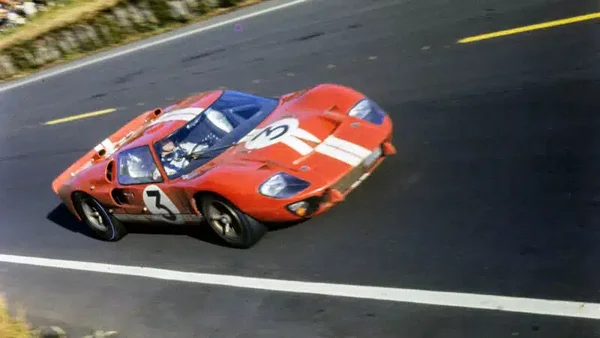 Une voiture de course Ford GT40 rouge avec des bandes blanches et le numéro 3, conduite sur une route pavée avec des marquages jaunes et blancs lors des 24 Heures du Mans.