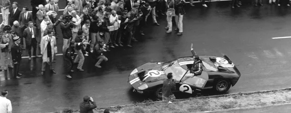 Une photographie en noir et blanc montrant une voiture de course vintage portant le numéro 2, défilant sur une piste mouillée devant une foule de spectateurs et de photographes.
