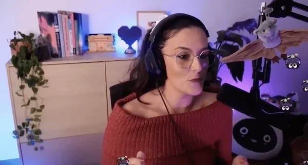 Ultia, femme aux cheveux foncés et lunettes, portant un casque et un pull rouge à épaules dénudées, assise devant un micro dans une pièce à éclairage violet avec une étagère de livres et des plantes.