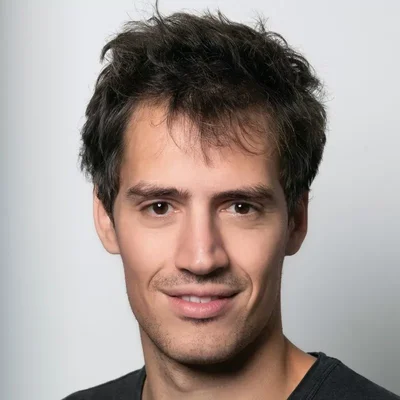 Portrait en gros plan d'Arthur Mensch, PDG de Mistral, jeune homme aux cheveux sombres décoiffés, yeux marron, légèrement barbe, souriant contre un fond gris clair