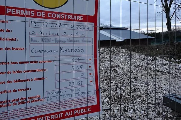Panneau "Permis de construire" installé sur une clôture grillagée d'un chantier à Noisiel, avec un bâtiment en construction et un sol en gravier.