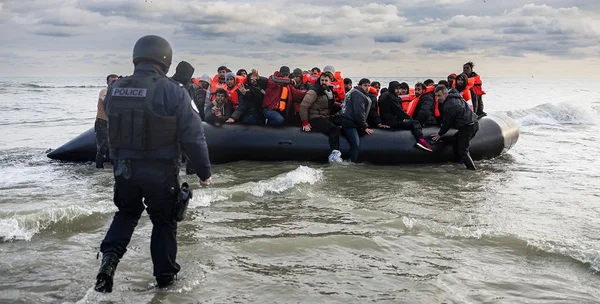 Un policier en équipement tactique se tient dans l'eau peu profonde face à une barque gonflable bondée de personnes équipées de gilets de sauvetage, qui approche de la rive.