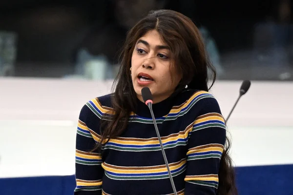 Rima Hassan, eurodéputée LFI, cheveux longs bruns et pull rayé, s'exprimant dans un microphone lors d'une prise de parole publique