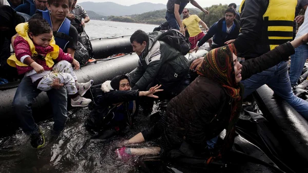 Un groupe de personnes, y compris un enfant, est débarqué d'une barque gonflable dans l'eau peu profonde, certains portant des gilets de sauvetage, sur les rives de l'Europe du Sud-Est.