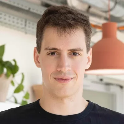 Portrait d'Arthur Mensch, co-fondateur et PDG de Mistral AI, jeune homme aux cheveux bruns courts, souriant légèrement, vêtu d'un t-shirt noir, dans un intérieur avec plante et lampe