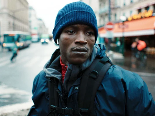 Portrait rapproché d'Abou Sangaré portant un bonnet et une veste bleus, dans une rue animée avec circulation et devantures floues en arrière-plan.