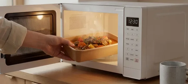 Une main retire un plat rectangulaire de viande et de légumes cuits d'un micro-ondes blanc ouvert, posé sur un plan de travail de cuisine.