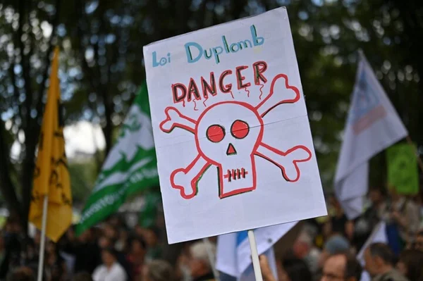 Pancarte dessinée à la main brandie dans une manifestation : une tête de mort surmontée du mot « DANGER » et l'inscription « Loi Duplomb », entourée de drapeaux et de manifestants