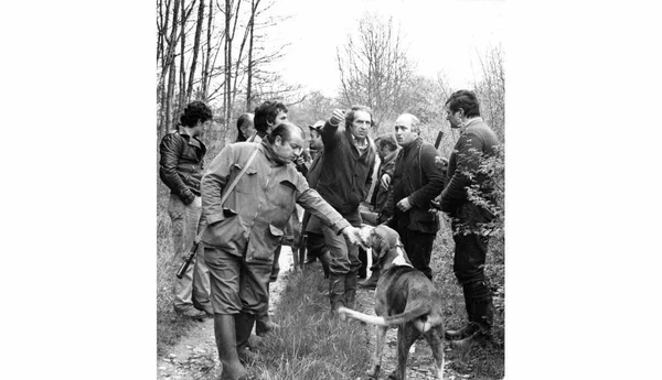 Photographie en noir et blanc d'un groupe d'adultes réunis sur un chemin forestier autour d'un chien, l'un gestuant et un autre caressant l'animal.