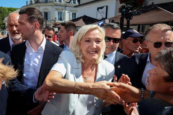 Marine Le Pen, souriante dans une veste claire, serre des mains lors d'un bain de foule près d'une terrasse de café, entourée de sympathisants et de membres de son service de sécurité, caméras visibles au second plan