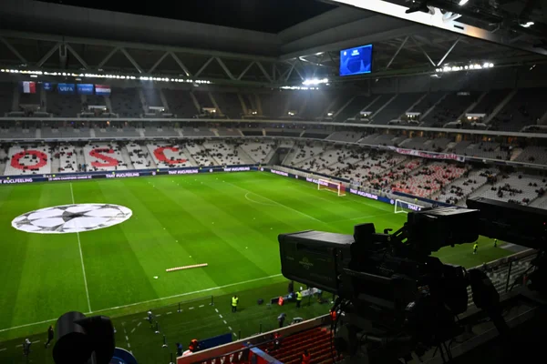 Vue plongeante depuis une caméra de retransmission sur un stade de football éclairé avant un match de Ligue des champions, pelouse ornée du logo UEFA, tribunes avec inscription PSG et spectateurs dispersés.