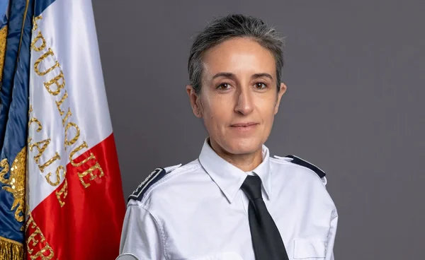 Portrait officiel de Céline Berthon, première femme à la tête de la DGSI, en chemise blanche de gendarmerie, cravate noire et épaulettes, devant le drapeau français sur fond gris.