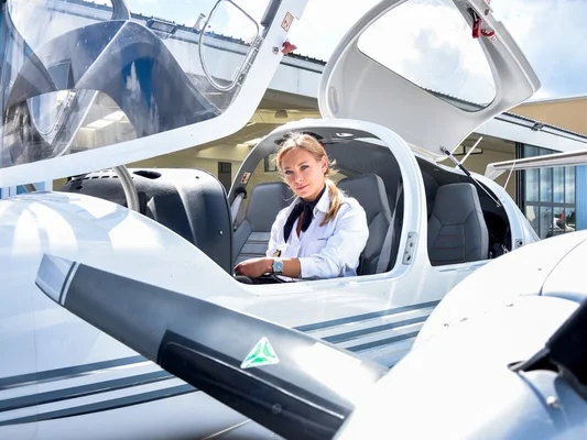 Une élève pilote en uniforme blanc et foulard sombre assise dans le cockpit d'un Diamond DA42 blanc à hélices, verrière ouverte, lors d'une formation multimoteur