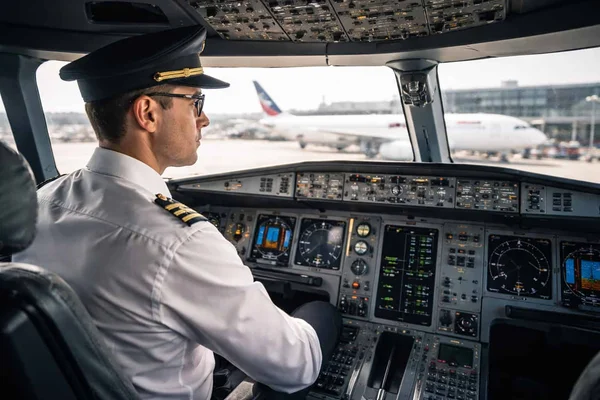 Pilote de ligne en uniforme blanc et casquette, assis aux commandes d'un cockpit d'avion de ligne, un autre appareil visible sur le tarmac à travers le pare-brise