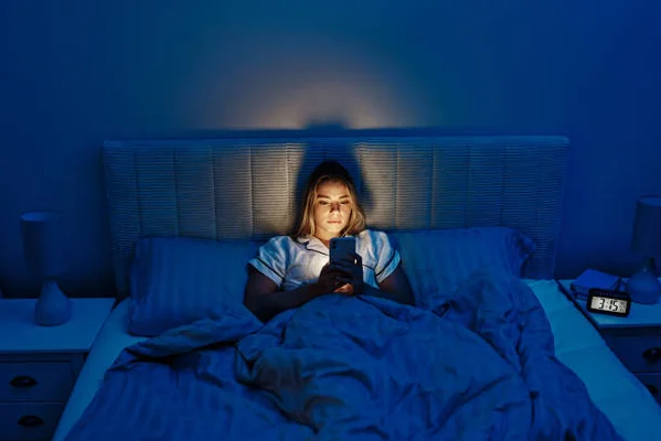 Jeune femme allongée dans un lit, dans une chambre éclairée en bleu, regardant son smartphone ; un réveil numérique sur la table de nuit indique 3 h 15.