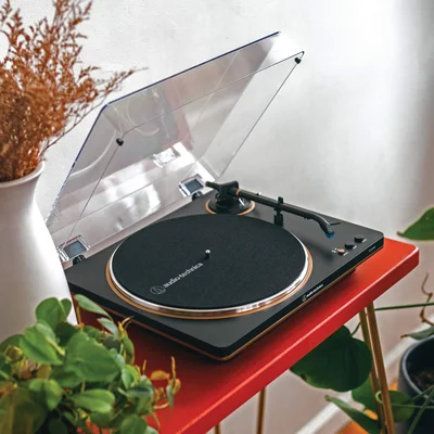 Platine vinyle Audio-Technica AT-LP70X capot transparent ouvert posée sur une table rouge à pieds dorés, entourée de plantes vertes et d'un vase blanc avec des fleurs séchées.