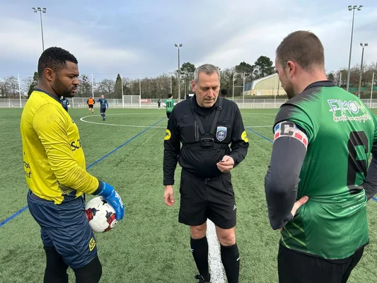 Un arbitre en tenue noire s'adressant à un gardien en maillot jaune tenant un ballon et à un joueur en maillot vert sur un terrain synthétique, avant le coup d'envoi d'un match de football en Sarthe