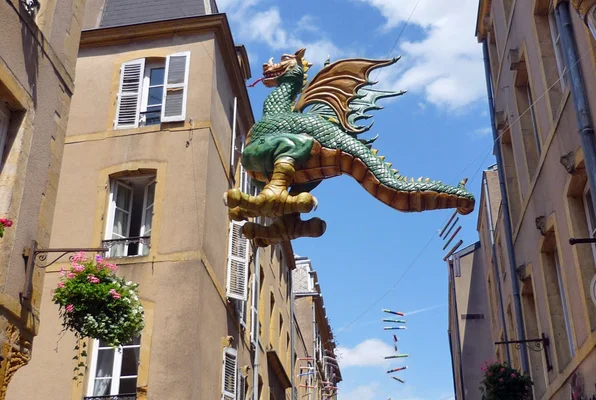 Le Graoully de Metz, grande sculpture verte suspendue entre deux immeubles de la rue Taison, au-dessus de la chaussée, avec des paniers de fleurs aux façades et un ciel bleu.
