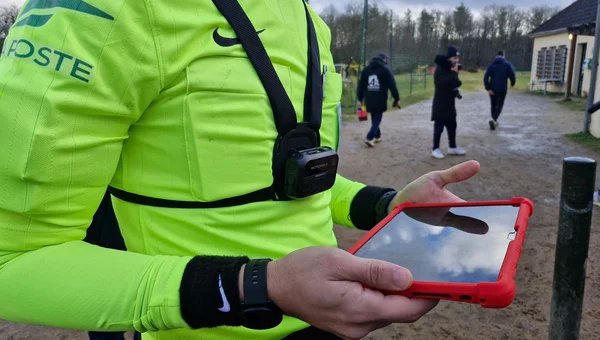 Un officiel en chasuble jaune fluo portant une caméra fixée sur un harnais et un bracelet au poignet, tenant une tablette dans une coque rouge sur un chemin de terre, des passants en arrière-plan
