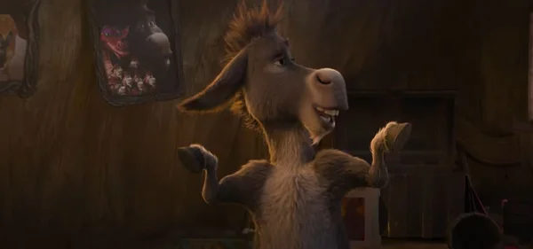 L'Âne, personnage de la saga Shrek, se tient dans une pièce faiblement éclairée avec des cadres photos accrochés au mur derrière lui.