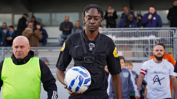 Un arbitre en tenue noire équipé d'une caméra embarquée tient un ballon bleu et blanc aux côtés d'un homme en chasuble jaune fluo sur la pelouse d'un terrain de football amateur en Bretagne, joueurs et spectateurs en arrière-plan