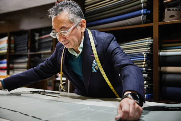 Un tailleur portant des lunettes et un blazer bleu marine, un mètre ruban jaune autour du cou, est penché sur une grande table de travail pour marquer ou couper du tissu dans un atelier de sur-mesure, avec des étagères de rouleaux de tissu en arrière-plan.
