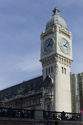Tour de l'horloge en pierre de la gare de Lyon, avec deux cadrans visibles et un dôme décoratif, surmontant un bâtiment de style classique sous un ciel partiellement nuageux.
