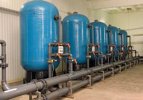 Rangée de grands cylindres bleus de filtration sous pression reliés par une tuyauterie grise et des vannes, installés dans un local technique d'usine de traitement des eaux