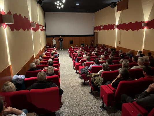 À Semur-en-Auxois, un public installé dans des fauteuils rouges écoute un intervenant debout devant un grand écran lors d'un ciné-débat associatif.