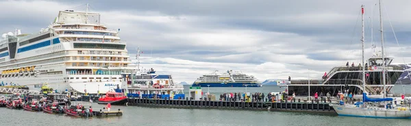 Vue panoramique du port de Svalbard avec un grand navire de croisière AIDA à quai à gauche, des passagers et de petits bateaux sur la jetée au centre, un navire bleu au loin et un voilier à droite sous un ciel couvert.