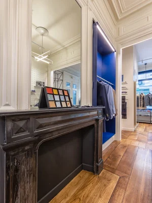 Intérieur d'un atelier de costume sur mesure à Lille : manteau de cheminée en bois foncé avec présentoir d'échantillons de tissus et miroir, alcôve de vêtements éclairée en bleu et parquet
