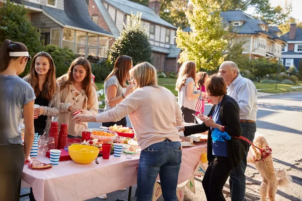 Des voisins de tous âges réunis autour d'une longue table lors de la Fête des voisins, avec boissons, collations et une femme tenant un petit chien en laisse.